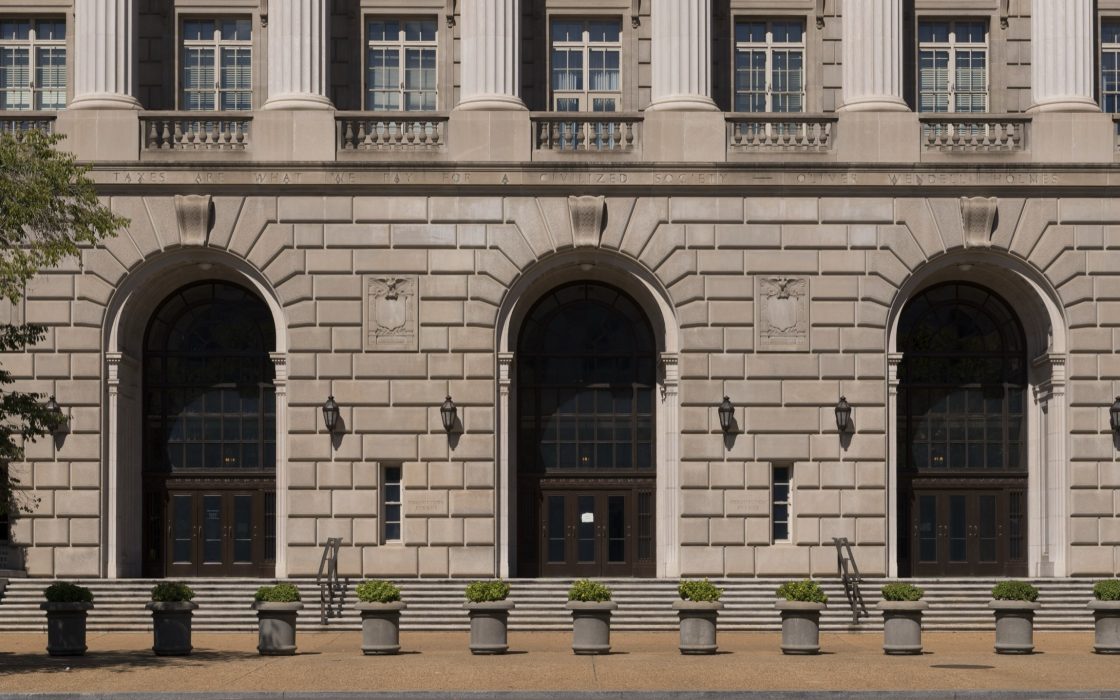 Limestone facade of the IRS headquarters building with three tall arched doorways and potted shrubs along the steps