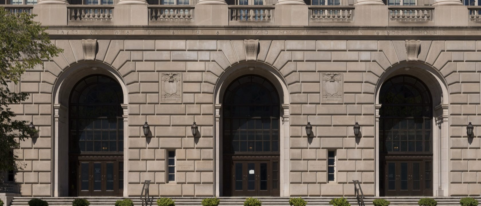 Limestone facade of the IRS headquarters building with three tall arched doorways and potted shrubs along the steps