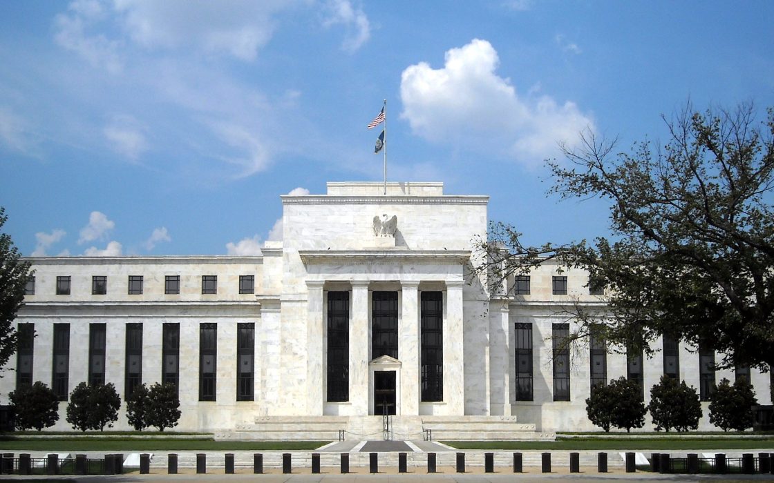 White marble facade of the Federal Reserve's Eccles Building under a blue sky, with a flag atop