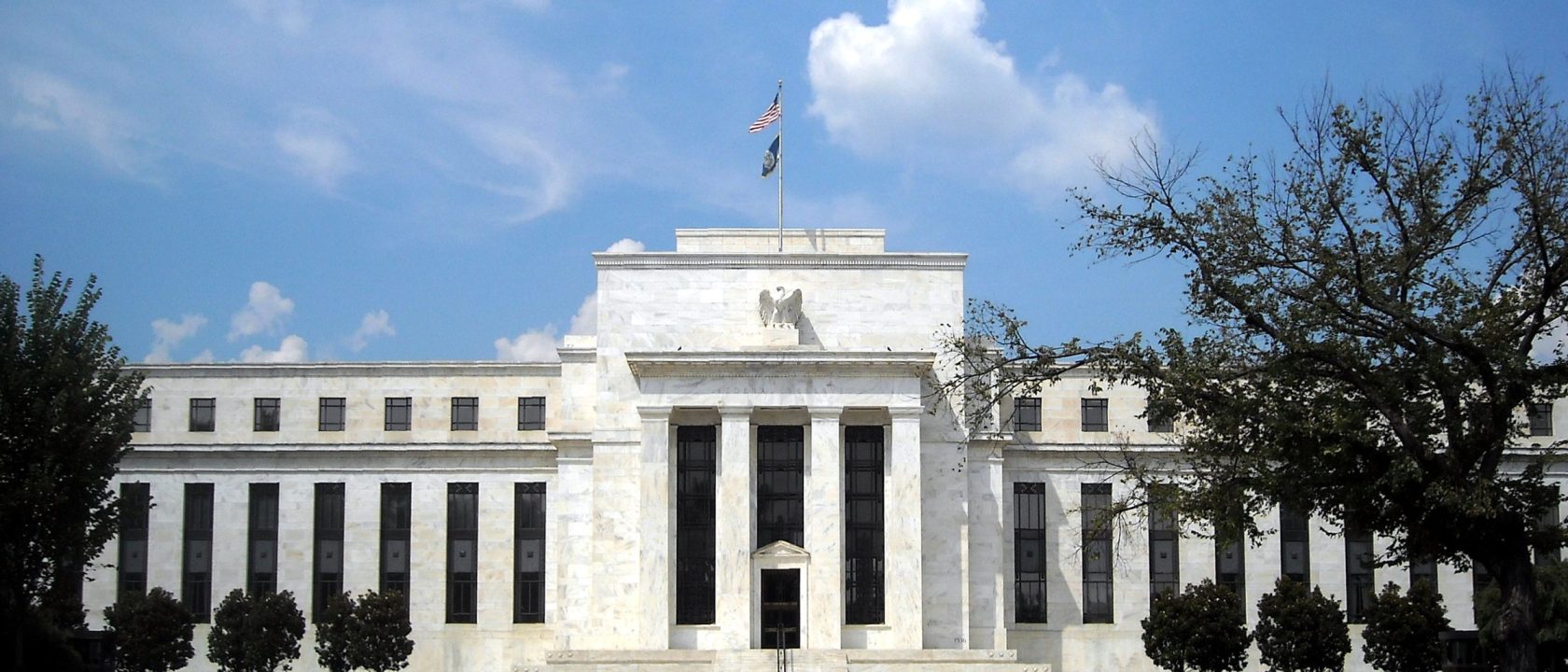 White marble facade of the Federal Reserve's Eccles Building under a blue sky, with a flag atop