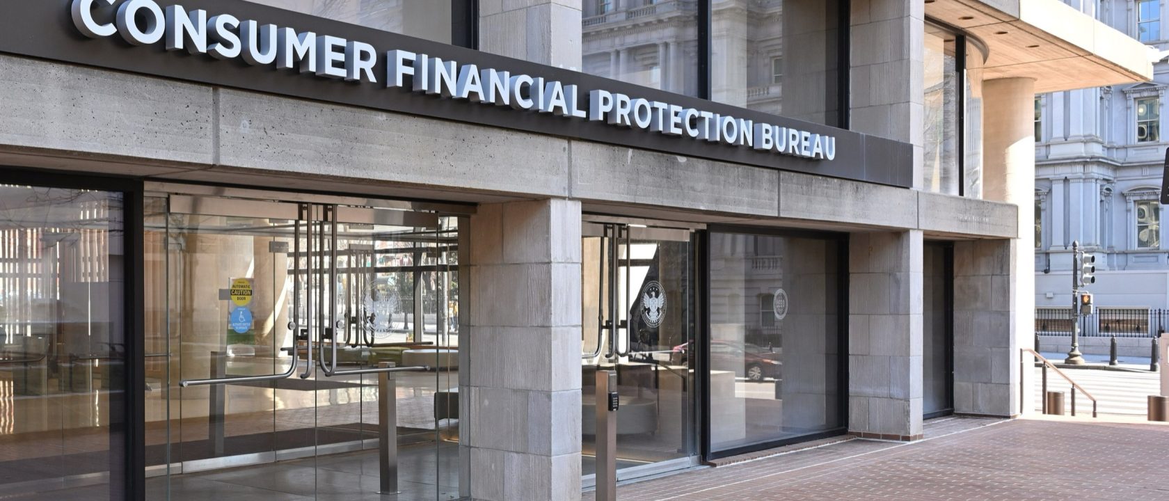 Glass entrance of the CFPB headquarters building with the bureau's name in raised letters above the doors