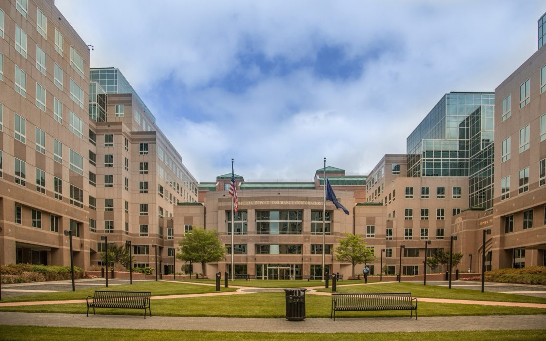 Tan brick office buildings around a lawn with benches and two flagpoles at the FDIC campus in Arlington, Virginia
