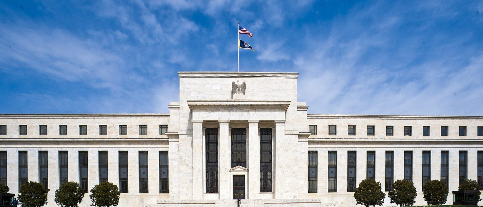 Front facade of the white marble Eccles Building with an American flag on top under a blue sky