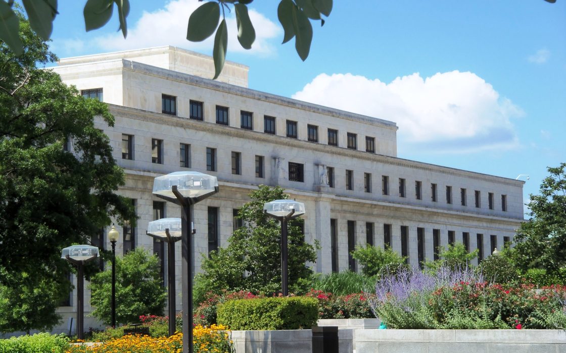 White marble Federal Reserve Eccles Building behind trees, flowers and street lamps under a blue sky