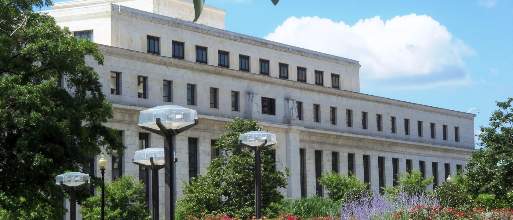 White marble Federal Reserve Eccles Building behind trees, flowers and street lamps under a blue sky