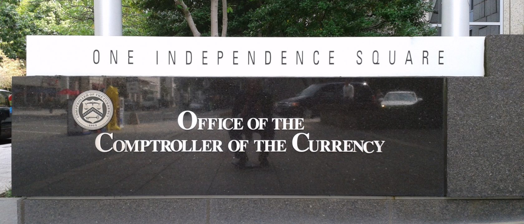 Polished black granite sign reading Office of the Comptroller of the Currency beneath a One Independence Square banner