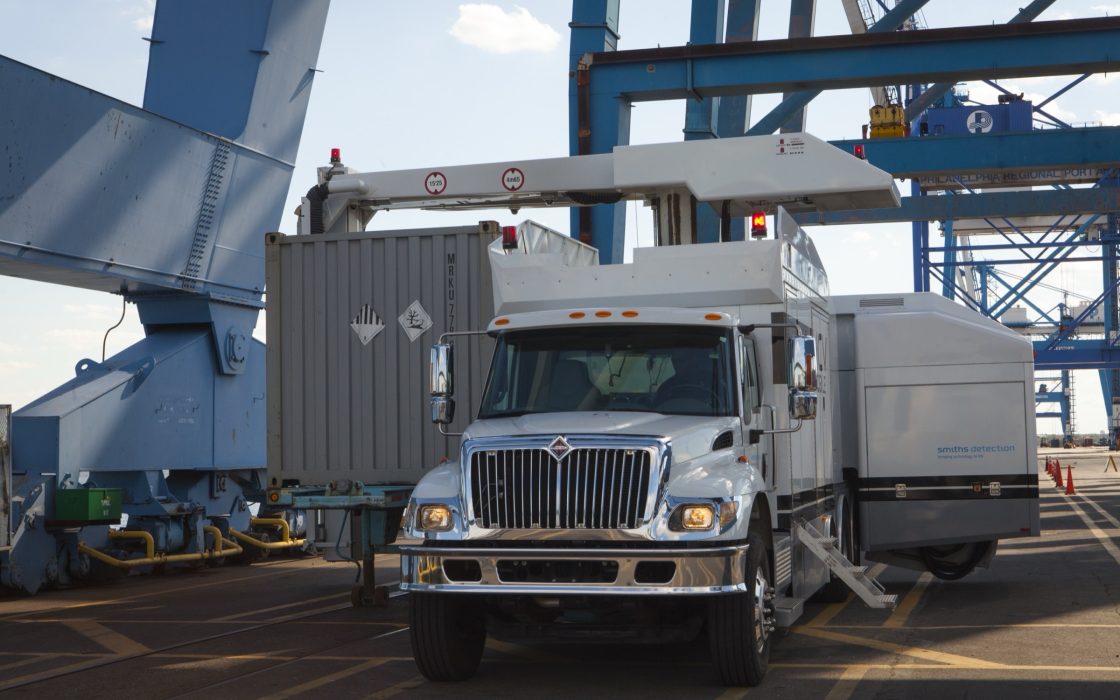 White CBP inspection truck with a scanning arm over a shipping container beneath blue port cranes