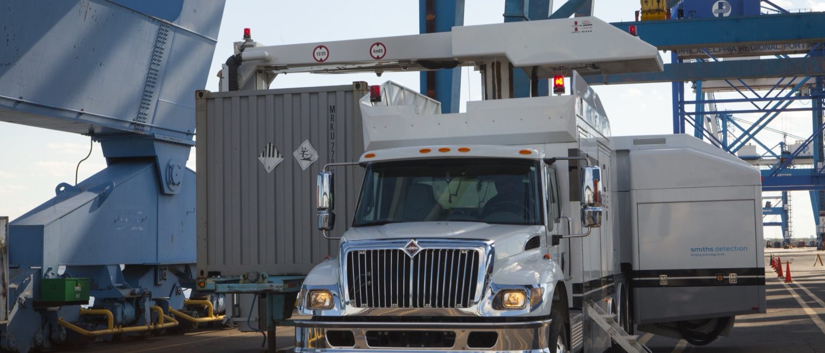 White CBP inspection truck with a scanning arm over a shipping container beneath blue port cranes
