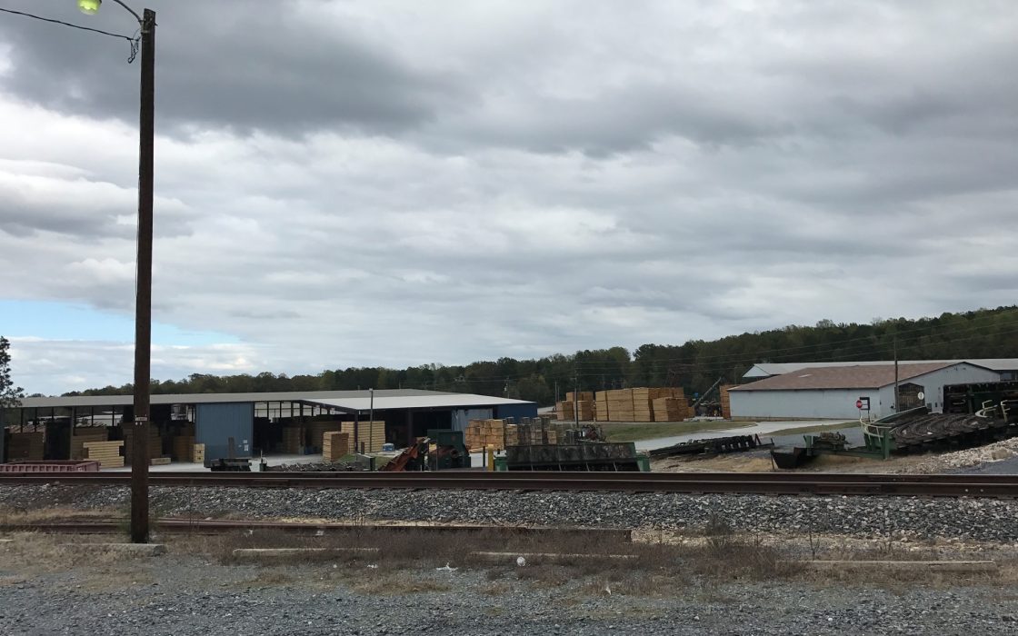 Lumber yard with stacks of cut boards and sheds behind a railroad track under a cloudy sky