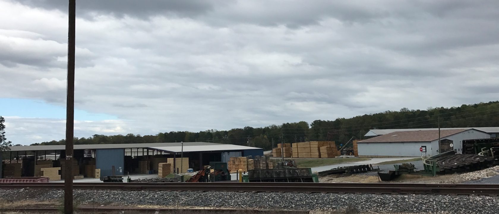 Lumber yard with stacks of cut boards and sheds behind a railroad track under a cloudy sky