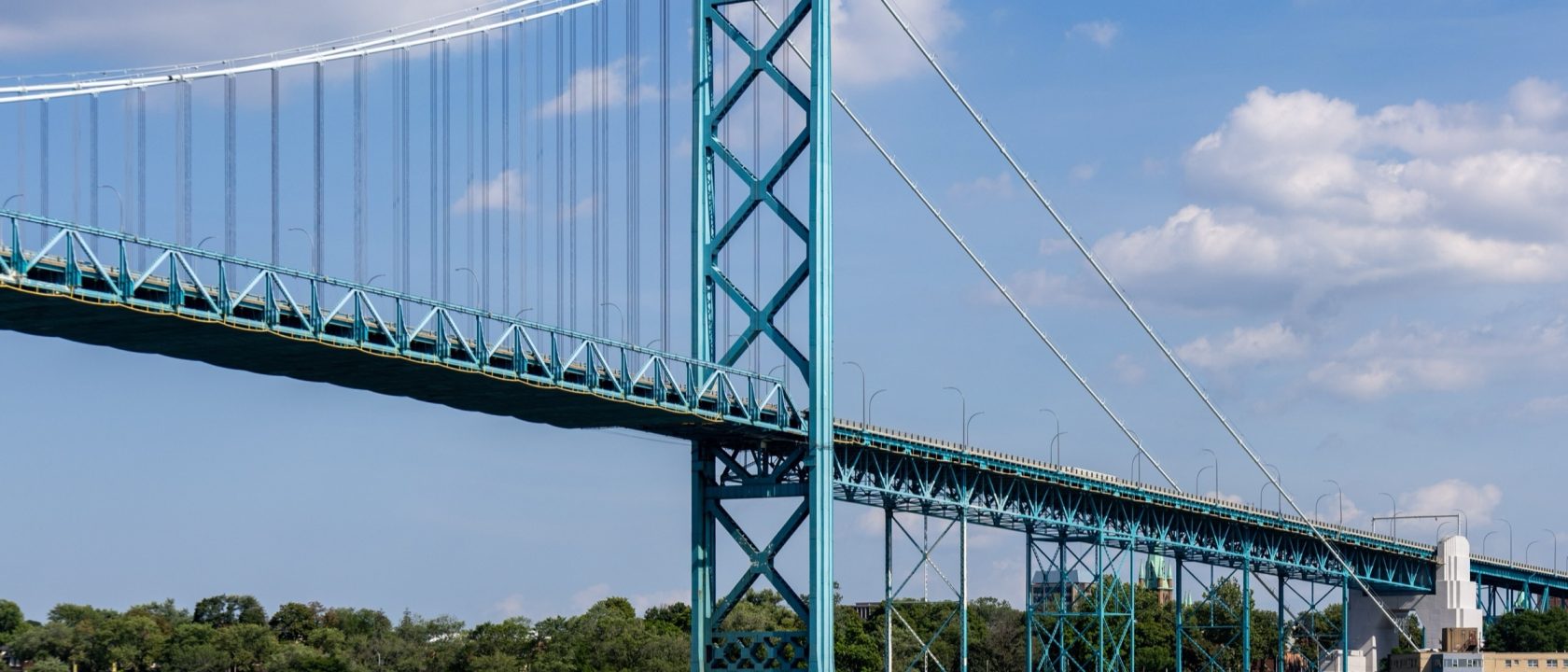 Teal suspension bridge spanning a river under a blue sky with clouds
